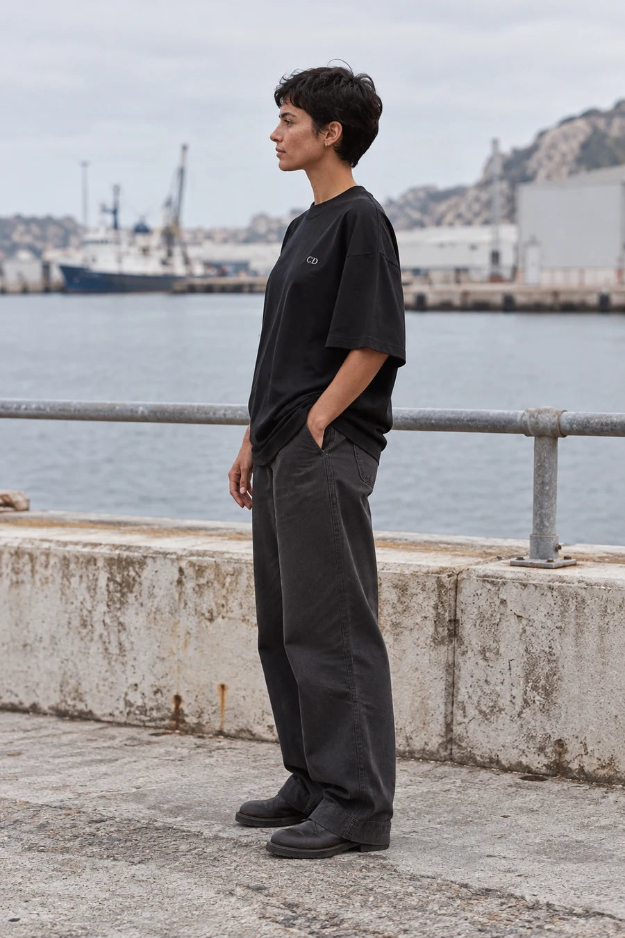 Model wearing a black Augusta CD T-shirt beside a harbour railing, shown from the side
